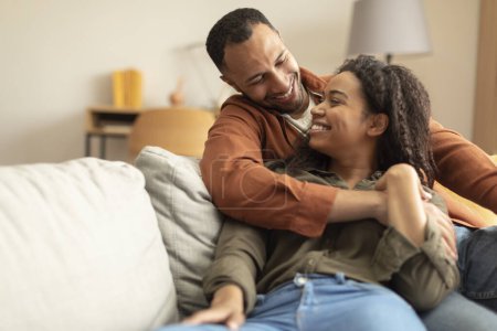 Téléchargez les photos : Loving black family embracing, looking at each other and smiling while relaxing on couch in living room, free space. Happy african american couple resting on sofa at home, spending weekend together - en image libre de droit