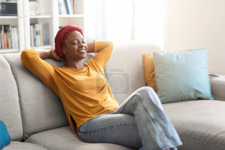 Foto de Relaxed peaceful happy young black lady in casual outfit with red turban on her head resting on couch at home, woman sitting on sofa with closed eyes and hands behind head, copy space - Imagen libre de derechos