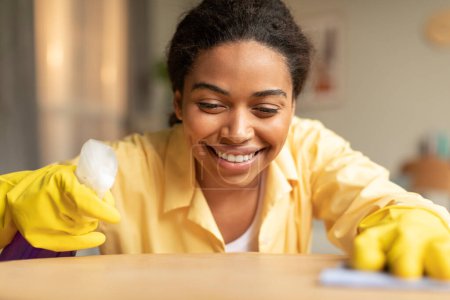 Foto de Happy black woman with sprayer detergent and rag cleaning table, lady wearing rubber gloves tidying home, closeup. Doing domestic chores - Imagen libre de derechos