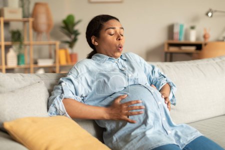Photo for Pregnancy pain. African american pregnant woman touching belly while sitting on couch at home, lady suffering painful contractions and making breathing exercises to relief ache - Royalty Free Image