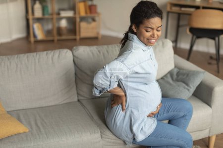 Photo for Sad black pregnant lady with big belly suffering from back pain, sitting on sofa in living room, free space. Motherhood, pregnancy problems, facial expressions, difficulties at last trimester - Royalty Free Image