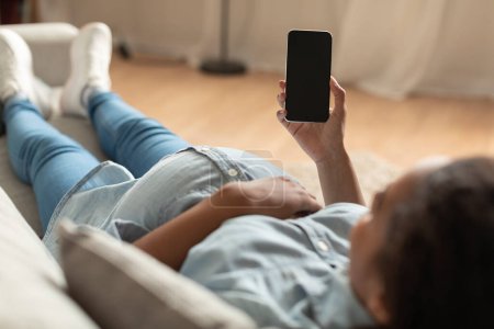 Foto de Black pregnant lady holding cellphone with blank screen, websurfing or reading maternity blog, lying on sofa at home. Unrecognizable woman using smartphone, mockup - Imagen libre de derechos