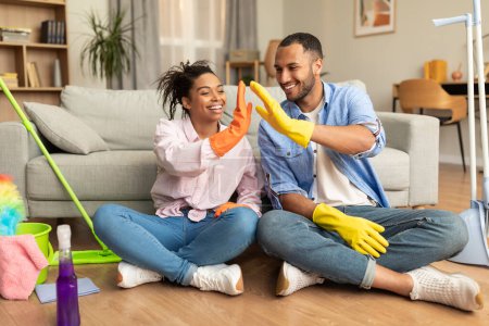 Foto de Fun cleaning together. Happy african american spouses in rubber gloves giving high five, sitting on floor in interior of living room with cleaning supplies nearby - Imagen libre de derechos