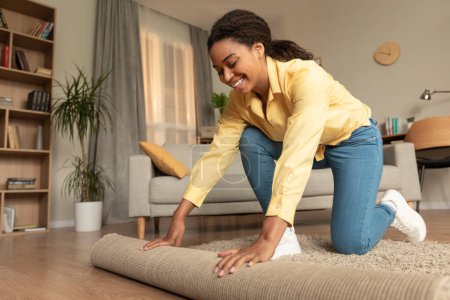 Foto de Young african american woman doing the housekeeping, bending down and rolling carpet in a living room, preparing to cleaning the floor, copy space - Imagen libre de derechos