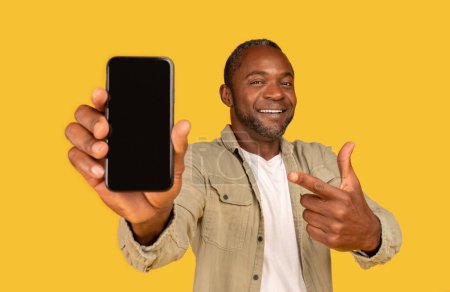 Photo for Cheerful middle aged african american male in casual show finger at smartphone with empty screen, recommending app, isolated on yellow background, studio. Blogger advice, new gadget, ad and offer - Royalty Free Image
