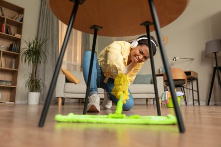 Téléchargez les photos : African american woman trying to mop floor under table, cleaning modern living room at home and listening to music in headphones. Low angle shot - en image libre de droit