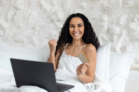 Téléchargez les photos : Glad arab millennial curly lady woke up, sitting on white bed with laptop, making victory and success gesture in bedroom interior. Good morning alone at home, work and business remote, great news - en image libre de droit