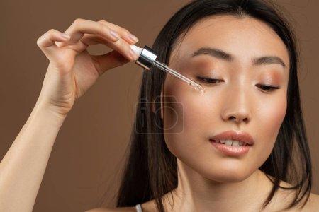Photo for Pretty young asian lady using nourishing face serum after shower, enjoying organic beauty product, standing on brown studio background, closeup shot - Royalty Free Image