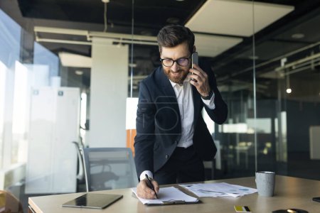 Foto de Corporate communication. Happy male manager working with papers and talking on cellphone, taking notes at financial documents, standing near workplace in office, free space - Imagen libre de derechos