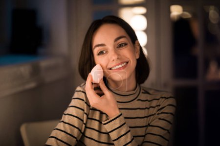 Téléchargez les photos : Cheerful smiling pretty brunette young lady with nice hairstyle applying makeup foundation at home, going out or testing brand new cosmetics, using sponge and looking at copy space - en image libre de droit