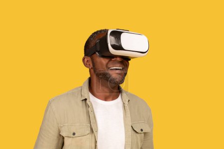Téléchargez les photos : Cheerful middle aged african american man gamer in casual and vr glasses enjoy online game and virtual world isolated on yellow background, studio. Entertainment and fun with device, ad and offer - en image libre de droit