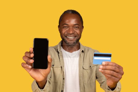 Foto de Cheerful mature black male show phone with empty screen, credit card to check bank financial account isolated on yellow background, studio. Recommendation gadget, app for online shopping and cashback - Imagen libre de derechos