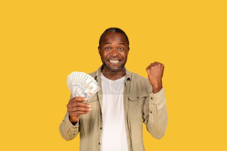 Foto de Glad excited adult african american male show a lot of money, cash, raises hand up, rejoices success, recommends investment, isolated on yellow background, studio. Financial savings, profit, win - Imagen libre de derechos