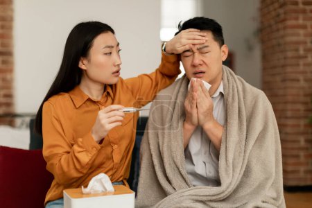 Foto de Asian woman treating sick husband, holding thermometer and touching man forehead, male having seasonal flu and cold, sitting on couch at home. Fever, influenza treatment concept - Imagen libre de derechos