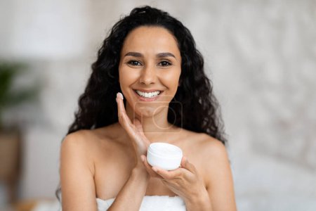 Téléchargez les photos : Portrait of cheerful middle eastern millennial curly lady in towel holding jar, applies cream on face in bedroom interior, close up. Beauty care and anti-aging treatments for skin, hydration, spa day - en image libre de droit