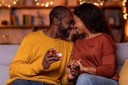 Téléchargez les photos : Emotional handsome bearded middle aged african american man holding his smiling happy girlfriend hand and beautiful engagement ring in red box, making marriage proposal, festive home interior - en image libre de droit