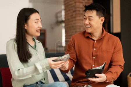 Foto de Wealthy korean mature husband giving money to his young wife, sitting together on sofa at home. Financial abundance family budget concept - Imagen libre de derechos