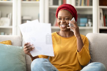Téléchargez les photos : Happy cheerful young black lady in casual outwear wearing red headscarf sitting on couch in living room, holding papers and talking on phone at home, having conversation with customer support - en image libre de droit