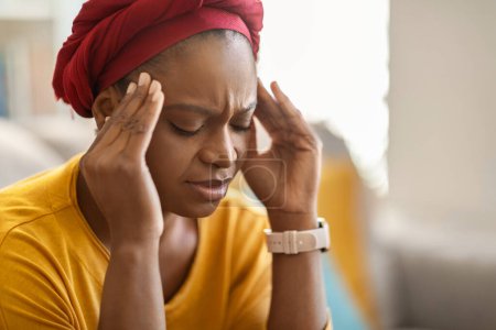 Photo for Exhausting migraine concept. Upset unhealthy young african american woman wearing red turban suffering from headache at home, sitting on couch, touching temples, closeup, copy space - Royalty Free Image