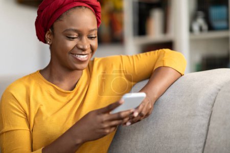 Foto de Positive cheerful relaxed pretty millennial black woman wearing red turban sitting on couch at home, using modern smartphone, chatting with guys on dating mobile app, copy space, closeup - Imagen libre de derechos