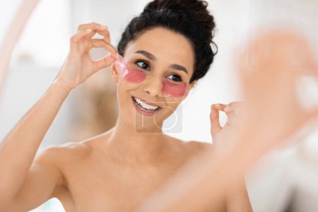 Téléchargez les photos : Portrait of happy arab millennial curly lady in towel apply eye patches looks in mirror, enjoy spare time in bedroom interior, close up. Skin and beauty care at home, anti-aging treatments and spa day - en image libre de droit