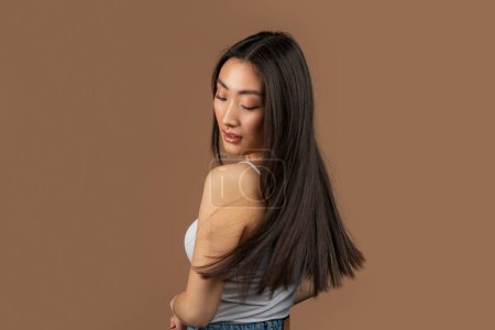 Foto de Young korean lady with perfect skin showing her long silky straight hair, posing on brown studio background, free space. Hair care and beauty treatments concept - Imagen libre de derechos