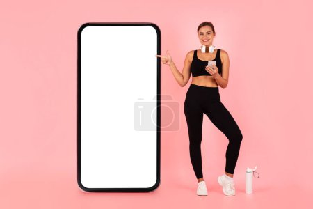 Téléchargez les photos : Happy Young Sporty Woman In Activewear Pointing At Big Blank Smartphone With White Screen, Smiling Athletic Female With Mobile Phone In Hands Advertising New Fitness App, Pink Background, Mockup - en image libre de droit