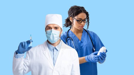 Photo for Multiracial medical team working on blue studio background, man and woman in white coats and protective medical face masks doctors holding syringe and jar with pills, panorama - Royalty Free Image
