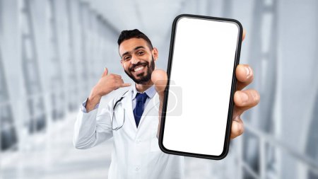 Foto de Handsome middle eastern male doctor showing blank smartphone and making call me gesture, arab therapist man in uniform holding phone with white screen while standing in hospital hall, collage, mockup - Imagen libre de derechos