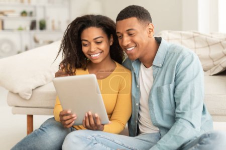 Foto de Technology for new normal at home. Glad millennial african american man and lady hug looking at digital tablet webcam in living room interior. Video call, meeting remote, chat and new app, covid-19 - Imagen libre de derechos