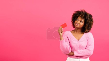 Téléchargez les photos : Thoughtful cheerful millennial african american curly lady shopaholic with credit card look at free space isolated on pink background, studio, panorama. Huge sale, online shopping, cashback and dream - en image libre de droit