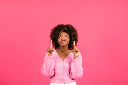 Foto de Happy pretty millennial african american curly female in casual show fingers up on blank space isolated on pink background, studio. Advice, recommendation, ad and offer, lifestyle and beauty blog - Imagen libre de derechos