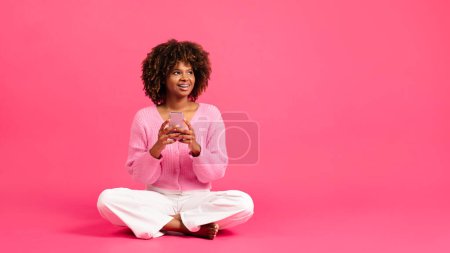 Téléchargez les photos : Glad inspired millennial african american curly lady with braces sit on floor, typing on phone, look at copy space isolated on pink background, studio. App for study, fashion blog, sale, ad and offer - en image libre de droit