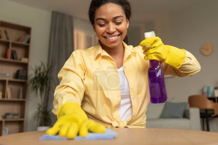 Foto de Cheerful african american woman in rubber gloves cleaning table in living room interior, using spray and duster cloth, free space. House-keeping concept - Imagen libre de derechos