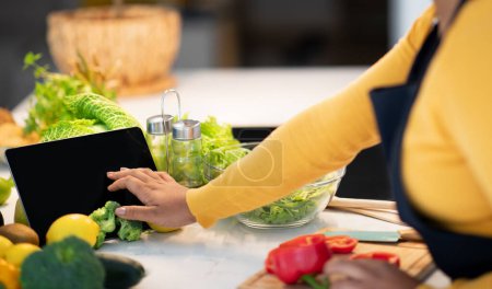 Téléchargez les photos : Young african american lady cook in apron use tablet with blank screen, watch food blog at table with organic vegetables, prepare salad in kitchen interior. App for new recipe, housework, diet at home - en image libre de droit
