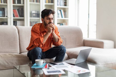 Photo for Pensive millennial middle eastern man suffering from burnout, sitting on couch, looking at copy space, working on laptop at home, overworking, looking for creative business solution - Royalty Free Image