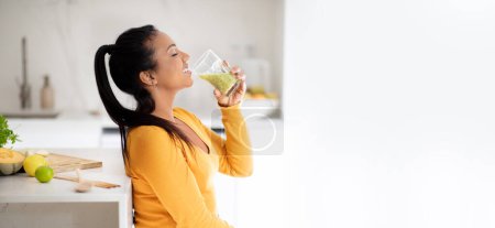 Photo for Glad young african american vegan woman at table with organic vegetables drink fresh smoothie at glass in minimalist kitchen interior, profile, panorama. Health, body care at home, diet, weight loss - Royalty Free Image
