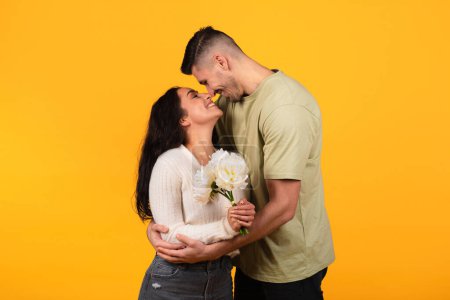 Photo for Happy millennial arabic guy hugging lady, gives bouquet of flowers, isolated on orange background, studio, free space. Couple enjoy love, romance and relationship, celebrate anniversary, date, holiday - Royalty Free Image