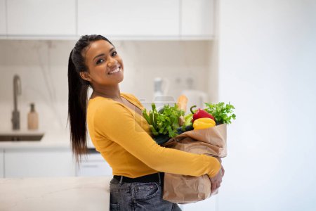Photo for Cheerful young african american lady holding paper bag with groceries in modern kitchen interior, free space. Products shopping, organic vegetables for weight loss, health care and food delivery - Royalty Free Image