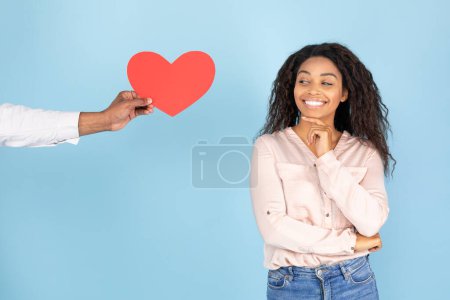 Photo for African american woman expressing happiness, man giving red heart shaped card to lady, female looking at it and smiling, standing over blue background - Royalty Free Image