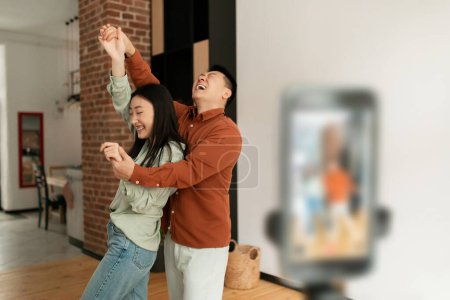 Téléchargez les photos : Playful asian spouses broadcasting or recording content while having fun together at home, vloggers dancing in front of smartphone camera, copy space - en image libre de droit