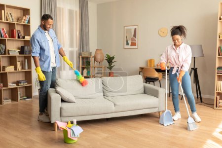 Téléchargez les photos : Shared house cleaning. Black man in rubber gloves and duster wiping dust while his wife sweeping the floor with broom and shovel in living room interior - en image libre de droit