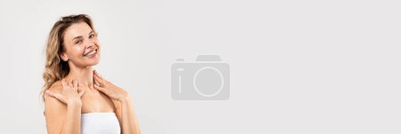 Téléchargez les photos : Spa Offer. Wide Banner With Beautiful Middle Aged Woman Wrapped In Bath Towel Posing On Light Grey Studio Background, Extended Shot Of Attractive Lady Touching Skin And Smiling At Camera, Copy Space - en image libre de droit