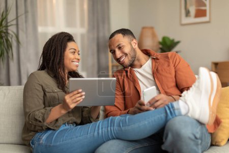Téléchargez les photos : Relaxed black spouses chilling together at home, happy man and woman using digital tablet and smartphone, resting on sofa. Entertainment and gadgets concept - en image libre de droit
