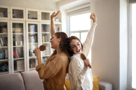 Téléchargez les photos : Happy excited european young superstar ladies have fun, sing song at imaginary microphone, enjoy free time in living room interior. Visit to friend, bachelorette party and dancing together at home - en image libre de droit