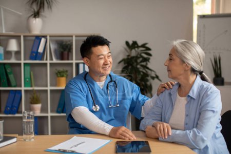Téléchargez les photos : Cheerful middle aged korean man doctor supports and advises, calms old european female patient in clinic office interior. Greeting, help, health care, treatment and visit to therapist, medical - en image libre de droit