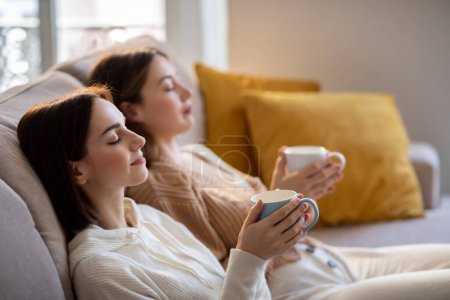 Photo for Calm european young women in casual with closed eyes relaxing, enjoy cups of tea, silence and comfort, sit on sofa in living room interior. Visit to friend, coffee break, rest and meditation at home - Royalty Free Image