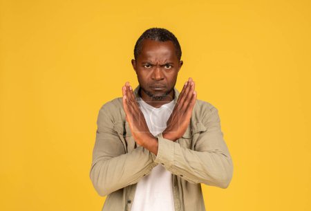 Téléchargez les photos : Angry confident sad middle aged african american man in casual crossed arms, doing stop sign isolated on yellow background, studio. Fight against discrimination, violence gesture and covid-19 virus - en image libre de droit