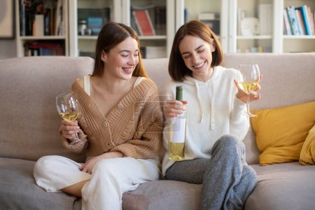 Photo for Laughing european young girlfriends with glasses and bottle of wine talking, enjoy spare time, sitting on sofa, have fun in living room interior. Visit to friend, bachelorette party together at home - Royalty Free Image