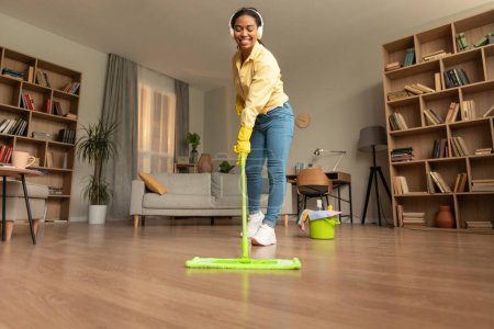 Téléchargez les photos : Young black woman washing floor in living room using mop and listening music in headphones, enjoying cleaning routine, living room interior, free space - en image libre de droit
